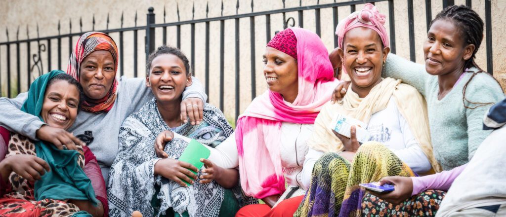 Six Ethiopian women sitting closely together outdoors, smiling warmly at the camera. They wear colorful traditional clothing including headscarves and wraps in teal, orange, pink, cream, and patterned fabrics. The women appear joyful and connected, with their arms around each other. A metal fence is visible in the background. The image represents community and togetherness, illustrating the Adera Foundation's work with families in Ethiopia.