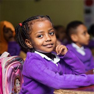 Young Ethiopian girl with braided hair and red hair ties, wearing a purple school sweater, smiling at camera while sitting at classroom desk.