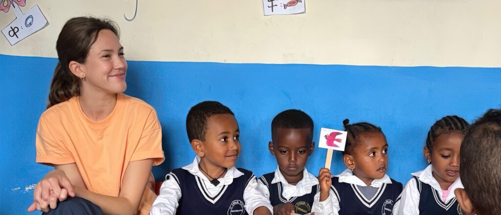 Woman in orange shirt sitting with five Ethiopian children in navy school uniforms in classroom with blue walls and educational posters