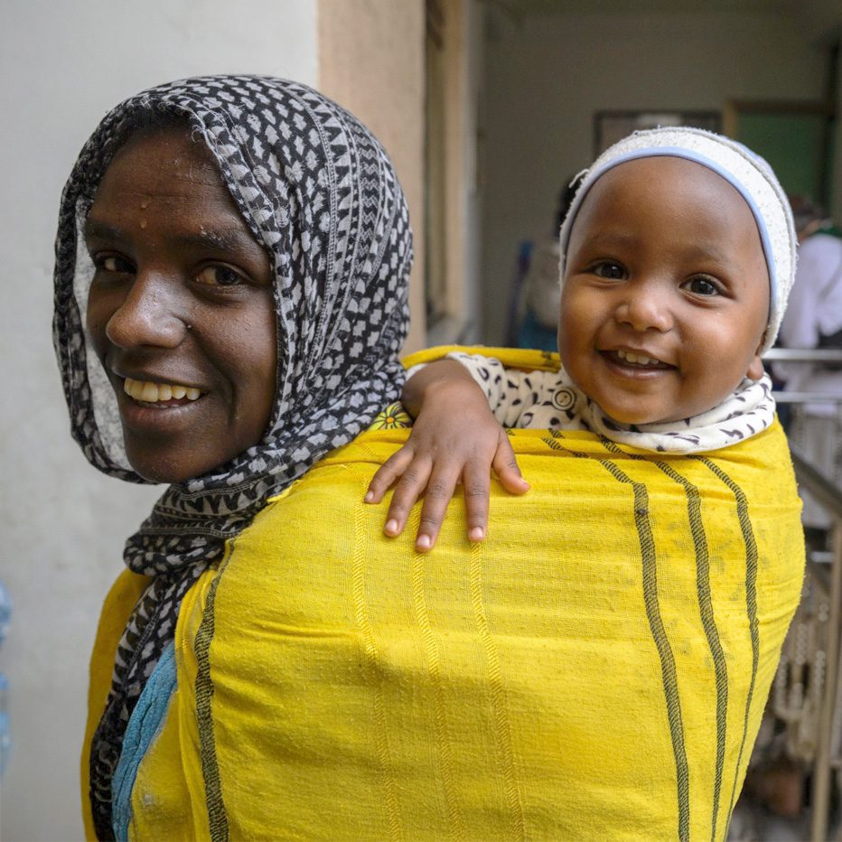 n Ethiopian mother carries her young child on her back using a bright yellow traditional cloth wrap with decorative striped borders. The mother wears a black and white patterned headscarf and has a warm, gentle smile. The child, wearing a white head covering, peeks over the mother's shoulder with a joyful expression and bright smile. The child's small hand rests on the mother's shoulder.