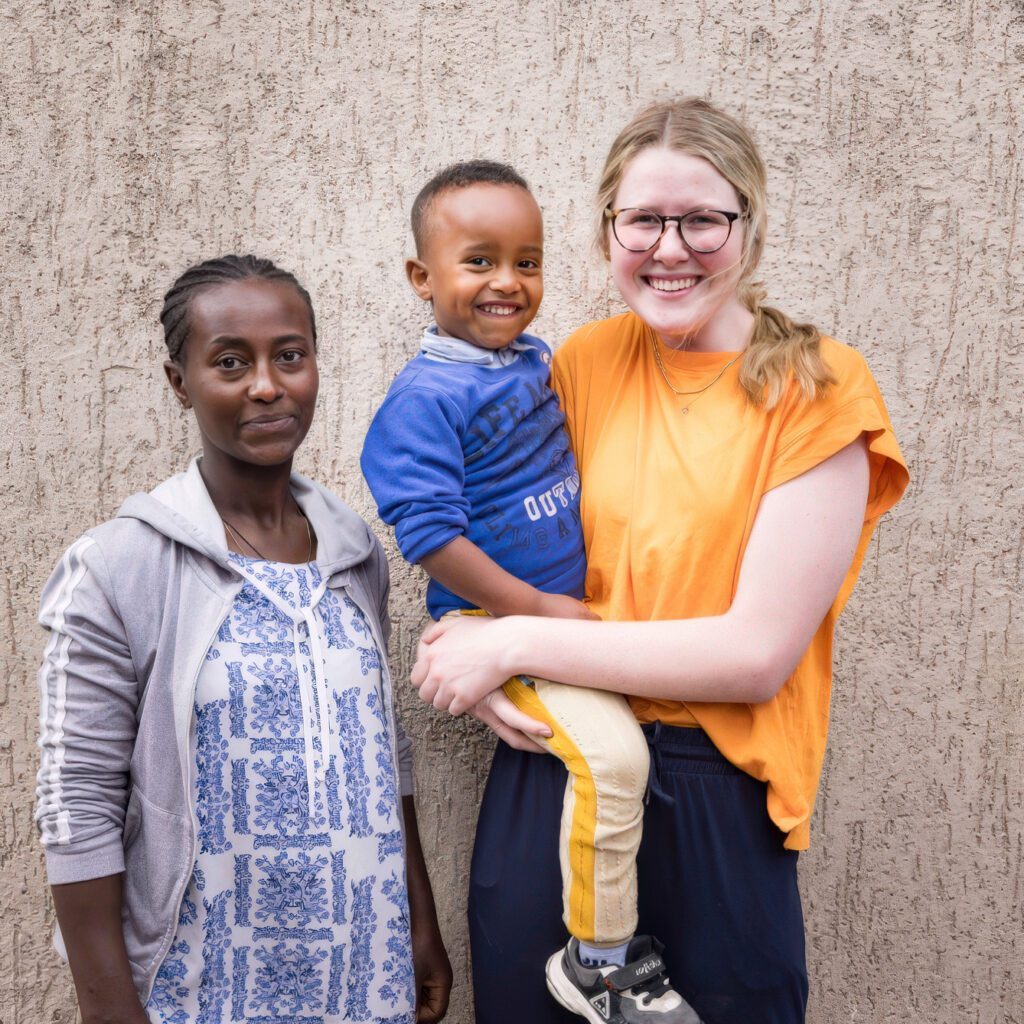 Ethiopian woman in gray hoodie stands beside blonde woman with glasses holding smiling Ethiopian boy in blue shirt - representing family sponsorship.