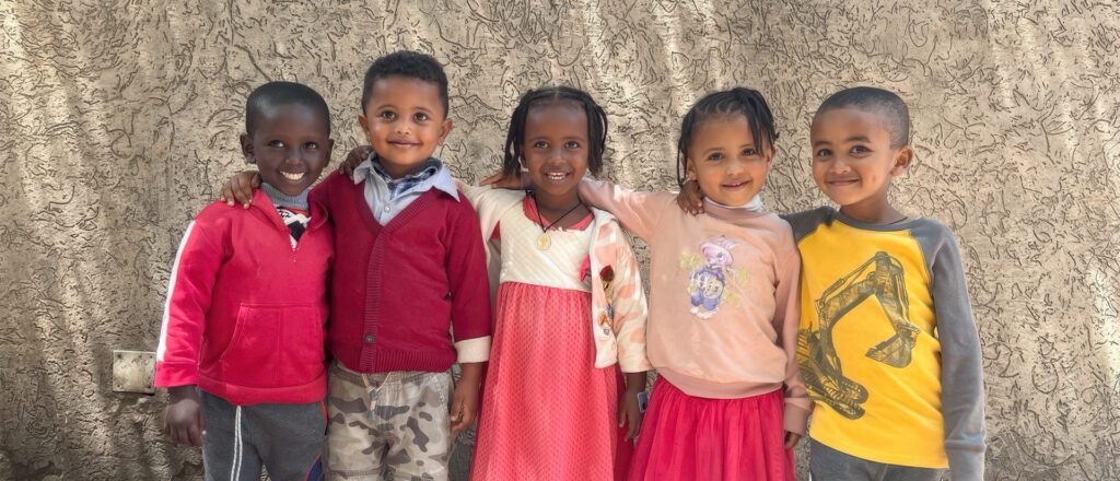 Five Ethiopian children standing arm-in-arm against a textured wall, wearing colorful clothing including red sweaters, a pink dress, and yellow shirt, all smiling warmly.