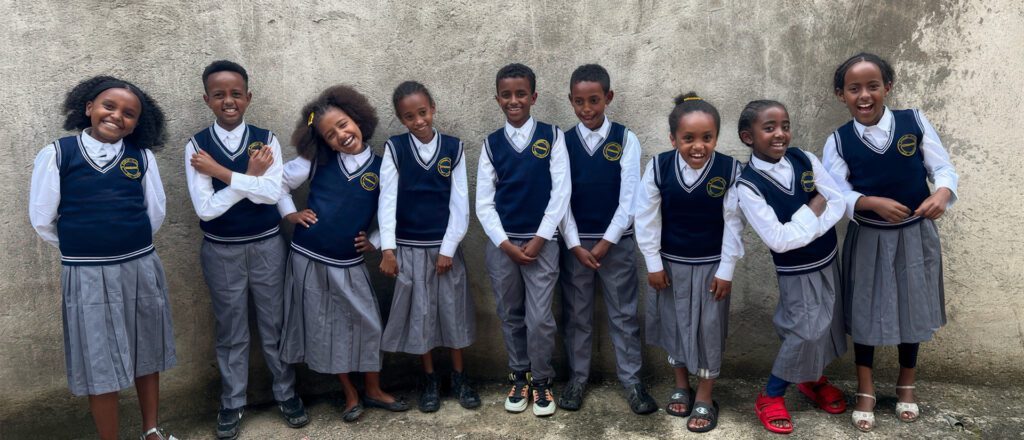 Nine Ethiopian school children in matching navy and white uniforms with gray skirts/pants, standing in a line against a concrete wall, all smiling brightly.