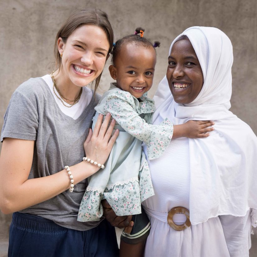 Three people pose together outdoors against a concrete wall background - a young Caucasian woman with brown hair wearing a gray v-neck shirt and pearl bracelet, a small Ethiopian toddler girl in a light green floral dress with her hair in small ponytails decorated with colorful hair ties, and an Ethiopian woman wearing a white headscarf and white clothing with a wooden bracelet.