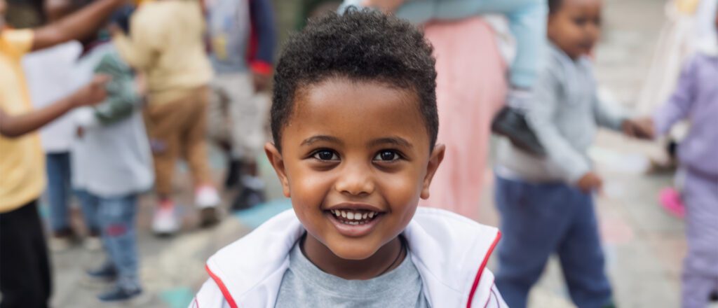 Smiling Ethiopian toddler boy in white and gray shirt with joyful expression, blurred crowd of people in colorful clothing in background.